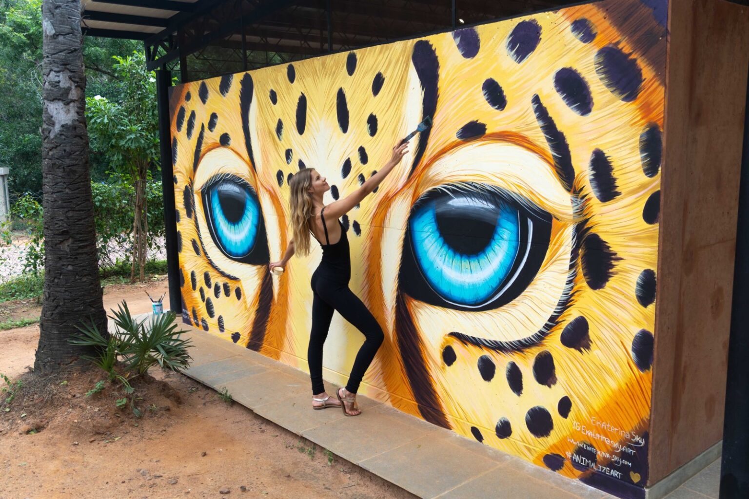 Ekaterina Sky in front of a mural of a leopard’s eyes, Animalize World Mural Tour